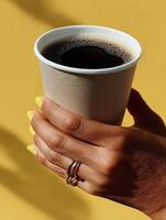 A minimalist close-up of a hand with yellow nail polish and silver rings holding a paper cup filled with black coffee. photo