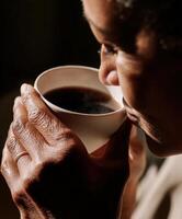 A close-up, intimate portrait of a woman holding a white ceramic cup with both hands, bringing it close to her lips as steam rises gently from the dark coffee. photo