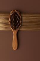 Minimalist top-down composition featuring a wooden paddle hairbrush with rounded bristles placed horizontally across a smooth sheet of silky blonde hair. photo