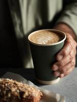 A minimalist coffee scene featuring a mans hand holding a dark green paper cup with a latte that has elegant leaf latte art on top. photo