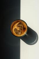 A top-down conceptual image of a glass of iced coffee with rich crema and large ice cubes, placed on a surface dramatically divided into light and shadow. photo