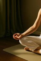 Partial view of a woman in white clothing sitting in lotus pose on a light yoga mat indoors. Her hand rests gently on her knee in a mudra gesture. photo