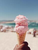 A close-up of a hand holding a pink strawberry ice cream cone against a bright, sunlit beach with blue sea and pale sky in the background. photo