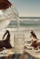 Close-up of a clear glass pitcher pouring cold water into a crystal glass filled with ice, placed on sandy beach with blurred ocean waves in the background. photo
