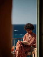 A woman sits in a wicker chair at a round table on a terrace by the sea, holding a book, with a cocktail glass next to her, against the backdrop of a sparkling ocean surface. photo