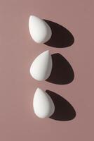 Minimalist flat lay photo of three white teardrop shaped makeup sponges beauty blenders arranged vertically with dramatic shadows on a muted dusty pink background.