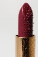 Macro close-up of a luxurious burgundy lipstick bullet covered in fresh water droplets, standing vertically in a shiny gold tube. photo