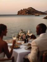 A romantic dinner scene with a couple enjoying wine and fine dining at a seaside table during sunset. The background features a tranquil sea with a small island and warm evening sky. photo