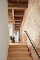 A modern industrial-style loft interior featuring a warm wooden staircase with clean lines, exposed red brick wall with aged plaster texture, and a rustic ceiling with visible wooden beams. photo