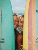 Two smiling surfer girls partially hidden behind colorful surfboards one turquoise and one yellow with a retro rainbow design standing side by side on a cloudy beach. photo