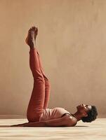 A woman with short natural hair practices a yoga pose lying on her back, holding her hips with both hands, legs extended upward with one bent at the knee. photo