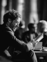 Black and white photograph of a stylish man with curly hair and glasses, wearing a formal suit, sitting in a cozy cafe while reading a book. photo