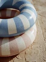 Two stacked inflatable swim rings in soft pastel colors, with wide cream and baby blue stripes, resting on a tiled poolside. photo