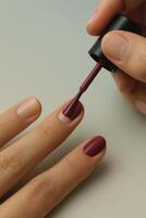 Macro shot of a hand applying deep burgundy nail polish to short, well-groomed nails. The brush is mid-stroke, just above the nail surface photo