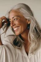 A middle-aged woman with long wavy silver hair smiles gently, tilting her head slightly and playing with a strand. She wears a flowing white blouse with soft texture and a drop-shaped earring. photo
