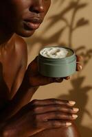 Close-up portrait of a dark-skinned woman holding an open jar of white body cream near her lips, against a warm beige background with soft botanical shadows. photo