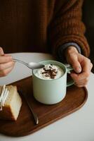 A cozy winter scene featuring a person in a chunky brown knitted sweater holding a mint green ceramic mug filled with whipped cream-topped hot chocolate, garnished with chocolate shavings. photo