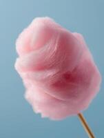 A close-up studio photograph of fluffy pink cotton candy on a wooden stick, isolated against a pastel blue background. photo