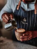 A detailed close-up of a barista in a striped apron pouring steamed milk from a stainless steel pitcher into a handcrafted ceramic cup, creating a perfect leaf-pattern latte art. photo