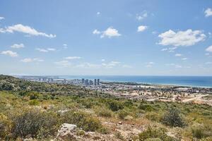 Expansive view of green hills and urban coastline meeting the Mediterranean, under a clear sky with scattered white clouds photo