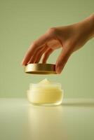 Close-up of a delicate hand gently lifting the golden lid off a frosted glass jar filled with soft yellow balm, placed on a smooth pastel green surface. photo
