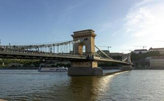 Side view of Chain Bridge in Budapest spanning the Danube River with suspension cables, stone pillars, and a tourist boat passing underneath photo