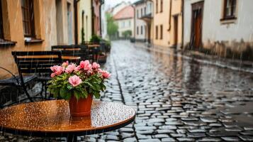 A quaint street scene with rain, a flower pot, and empty cafe tables on cobblestones. photo