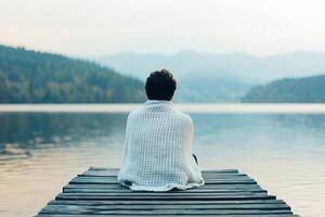 A person sitting on a dock, wrapped in a blanket, overlooking a tranquil lake. photo