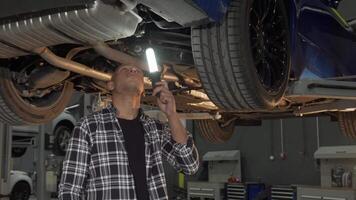 Handsome man inspecting lifted car with a flashlight in a modern workshop video