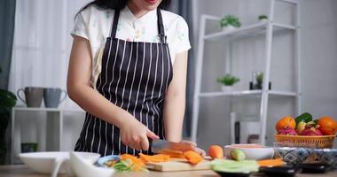 Handheld low angle view shot, Beautiful Asian woman cutting carrots with knife on wooden cutting board. Woman preparing fresh vegetables to make salad rolls. video
