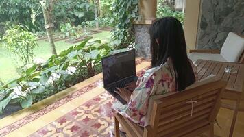 A woman enjoys working on her laptop in a serene outdoor setting, surrounded by lush greenery and a tranquil garden. Focus on creating while staying connected. video