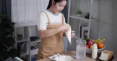 Handheld high angle view shot, Beautiful Asian woman scooping whipped cream into a piping bag to prepare to make dessert on the table at home. video