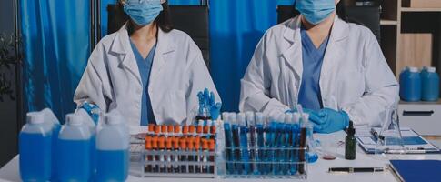 Two people in lab coats and masks standing behind a table with bottles and other supplies photo