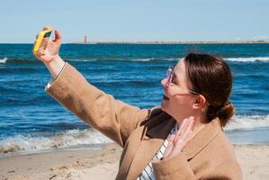 Caucasian young female enjoying sunny beach day takes selfie with yellow camera against vibrant blue ocean and lighthouse in the distance. Digital nomad lifestyle. Remote work photo