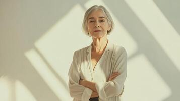 Mature caucasian woman in white blouse stands confidently in sunlit room with crossed arms and neutral expression, embracing natural light and shadow patterns on the wall photo
