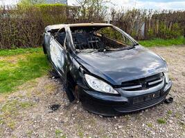 A completely burned-out black sedan with extensive fire and impact damage sits abandoned on a rocky patch beside a hedgerow in a rural area photo