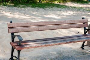 Weathered wooden bench on sandy pathway with elegant iron detailing in sunlit park surrounded by lush greenery and shadows creating serenity. Digital nomad lifestyle. Remote work photo