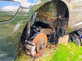 Close-up of a rusted wheel hub and suspension of a burned car with visible fire damage, corrosion, and exposed metal components on a grassy patch photo