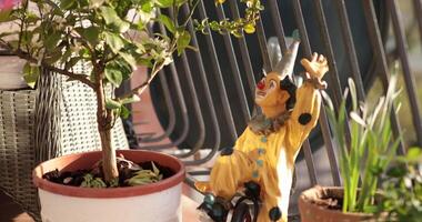 Bright mosaic flower pot stands out filled with lush greenery and decorative elements. Clown statue adds charm to balcony sunny ambiance, creating a delightful outdoor escape. video