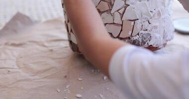 Person is carefully applying pieces of broken tiles to a flower pot, creating an artistic mosaic design. The workspace is bright and filled with creative energy, showcasing an engaging DIY project. video