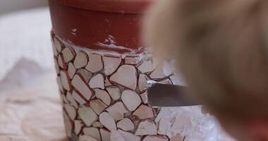 Process of designing broken tile mosaic flower pot. Person decorate flower pot with mosaics applying putty with spatula. DIY craft project for garden decorations video