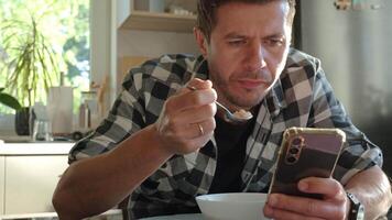 Man eating oatmeal porridge from white bowl while using smartphone. Male looking at phone during breakfast. Concept of social media addiction, distraction, and digital dependency video