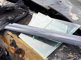 Broken window glass from a car resting on a burned and rusted door frame, with visible soot, melted rubber seals, and fire-damaged metal components photo