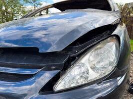 Close-up of a damaged black car front with a broken hood and intact headlight, reflecting the sky and trees in a rural outdoor setting after a fire photo