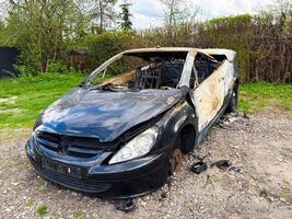 Severely burned black car with missing wheels and extensive fire damage, abandoned in a grassy field surrounded by trees and shrubs under a partly cloudy sky photo