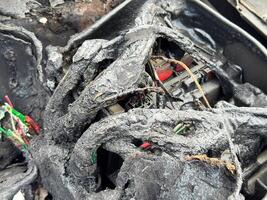 Burned car fuse box and wiring with melted insulation, scorched plastic housing, and exposed colored wires after extensive fire damage in the engine compartment photo