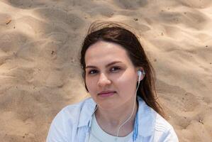 Young caucasian female with earphones at beachside, engaged in relaxing moment, wearing casual attire, surrounded by sandy shores. Digital nomad lifestyle. Remote work photo