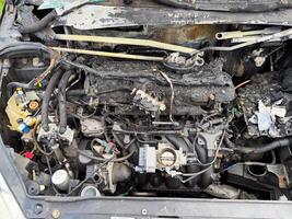 Burned car engine compartment showing fire-damaged engine block, melted wiring, scorched hoses, and exposed intake manifold with severe thermal destruction photo