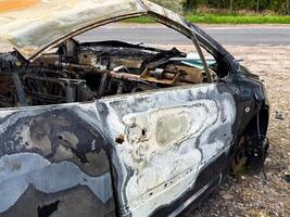 Side view of a completely burned car showing warped and blistered paint, missing interior, and rusted components after an intense fire incident near a roadside photo