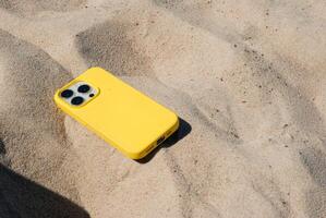 Bright yellow smartphone case on sandy beach under clear blue sky showcasing summer technology vibes in coastal outdoor setting. Digital nomad lifestyle. Remote work photo